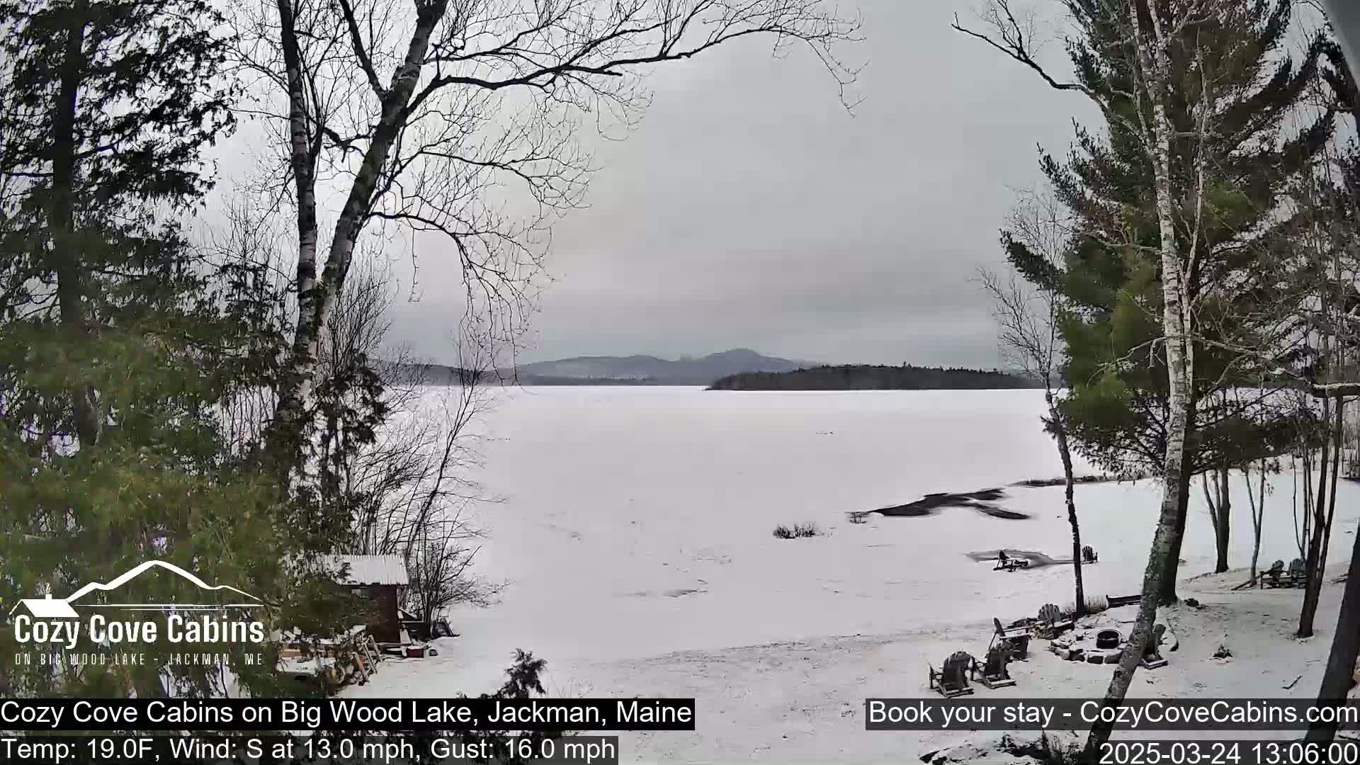 A snow-covered frozen lake is seen from a snowy shoreline with leafless trees and a small cabin, under an overcast sky with a south wind.