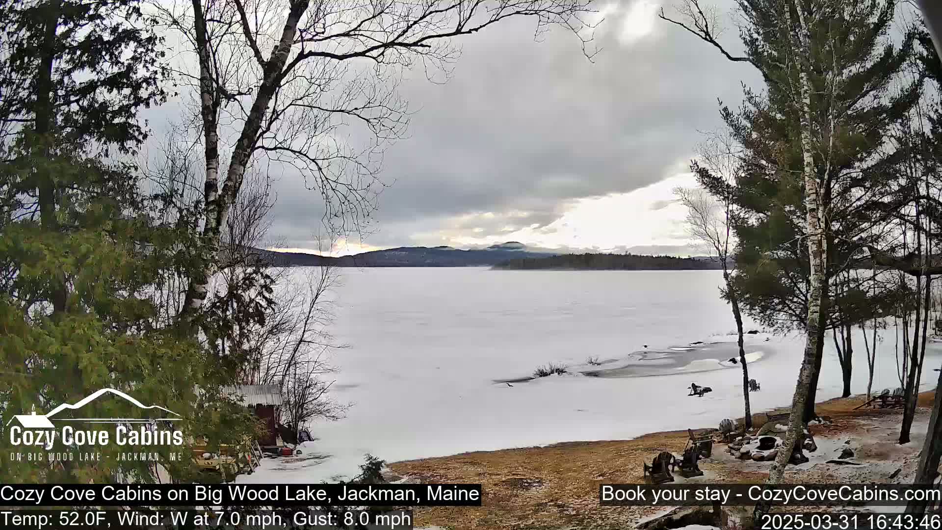 A partially frozen lake is seen from a lakeside cabin with leafless trees and a cloudy sky.