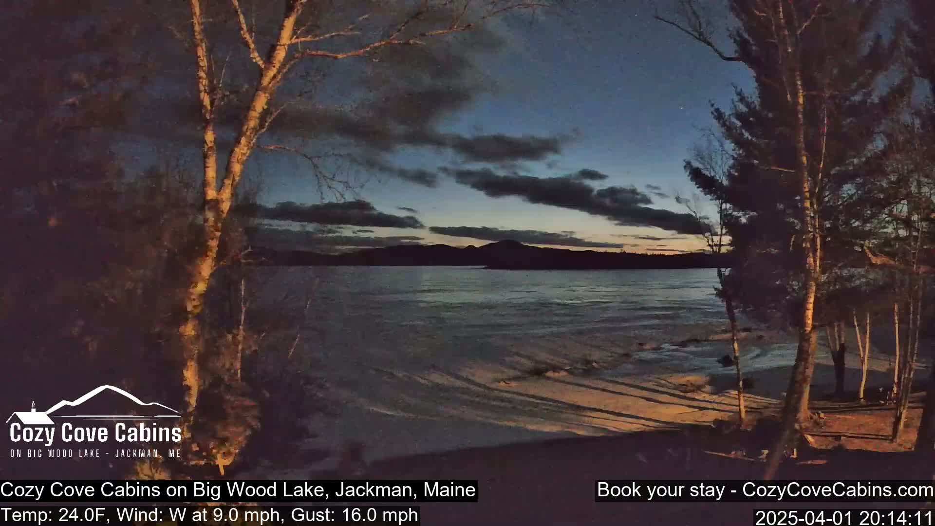 A twilight view of a lake with a partially frozen shoreline, under a partly cloudy sky, with trees in the foreground.