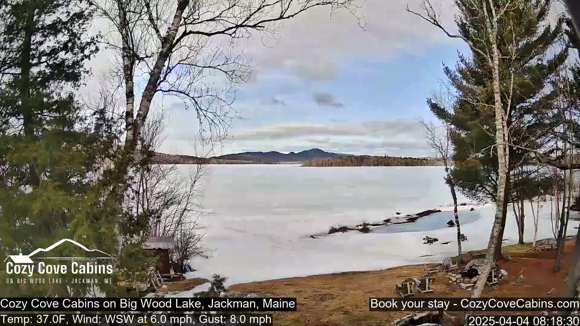 A partly cloudy day reveals a frozen lake with snow-covered banks, framed by leafless trees.