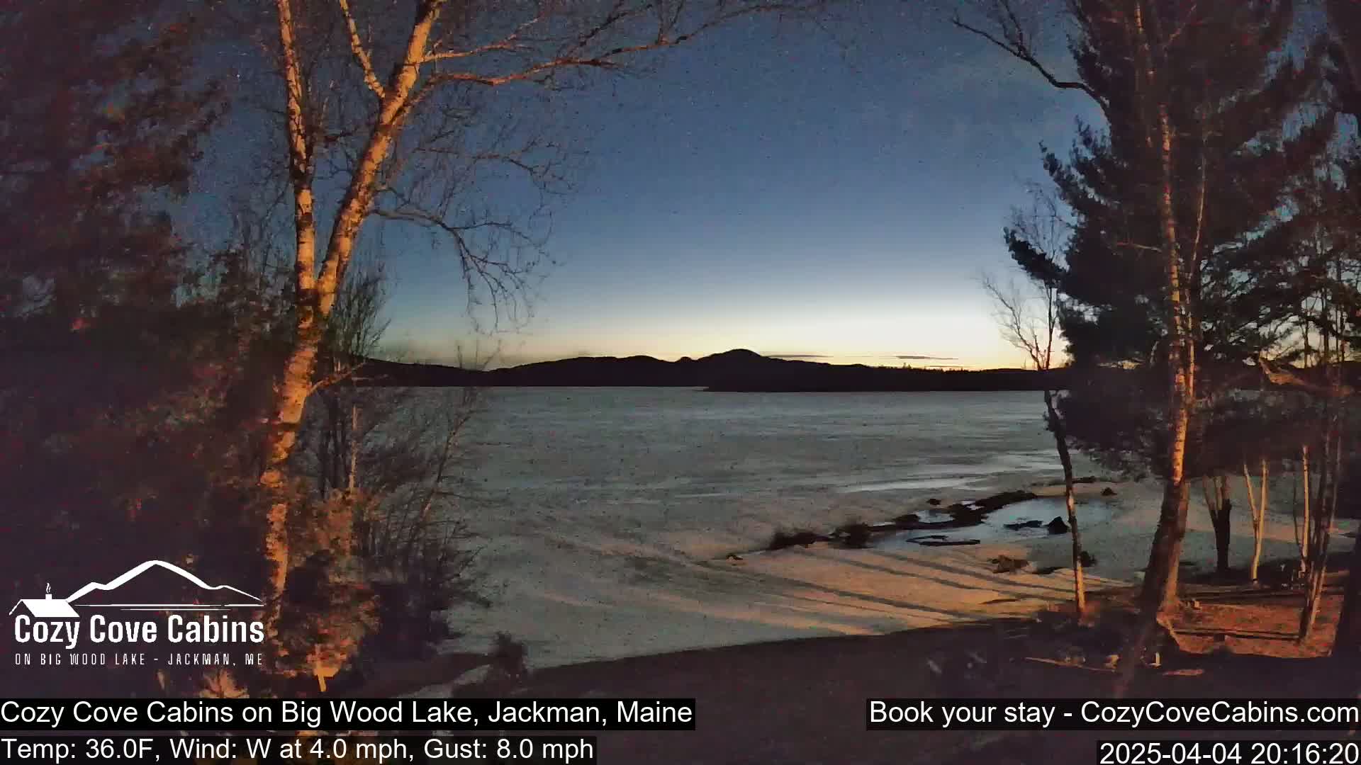 A partly frozen lake is seen at twilight, with bare trees in the foreground and mountains in the background, under a clear, dark blue sky.