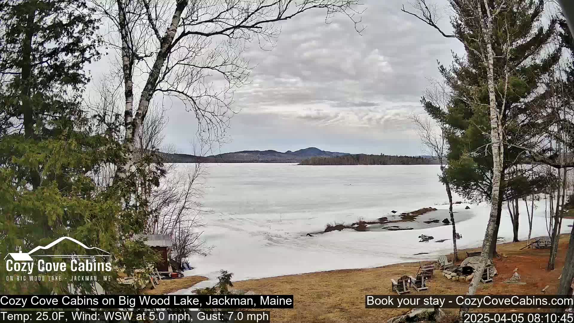 A partially frozen lake with snow-covered banks is viewed from a lakeside property under a cloudy sky.