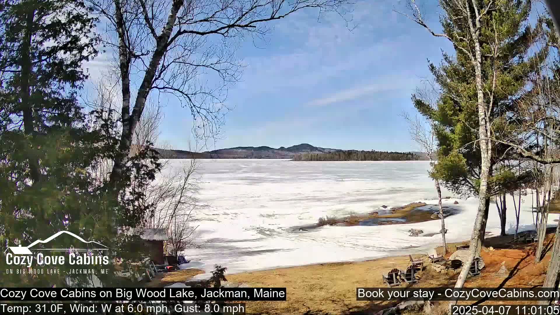 A partly frozen lake is visible from a lakeside vantage point under a mostly sunny sky, with bare trees in the foreground and distant hills in the background.