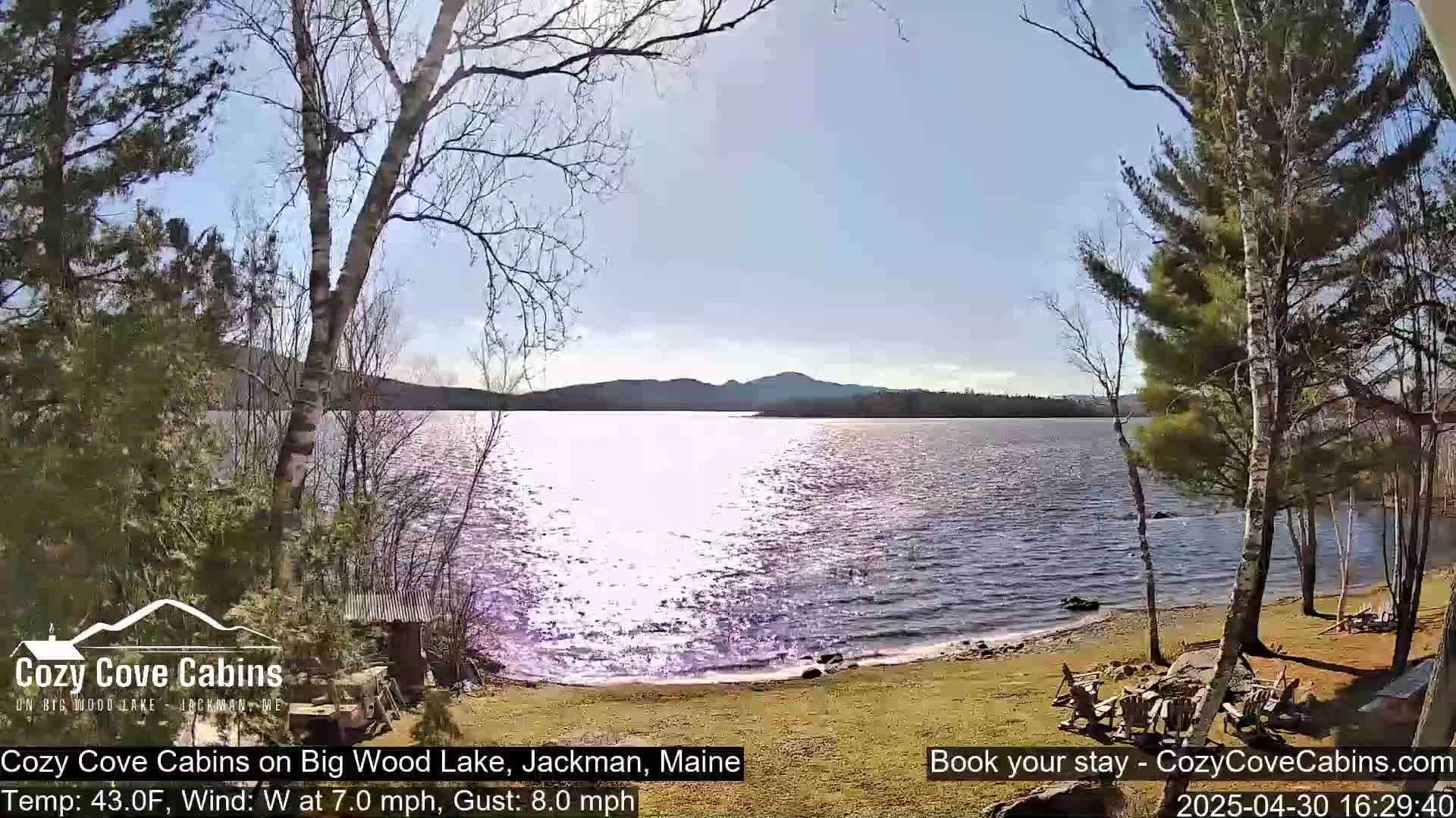 A sunny day with a 7 mph wind shows a view of a lake with a sandy shore, trees, and a small structure.