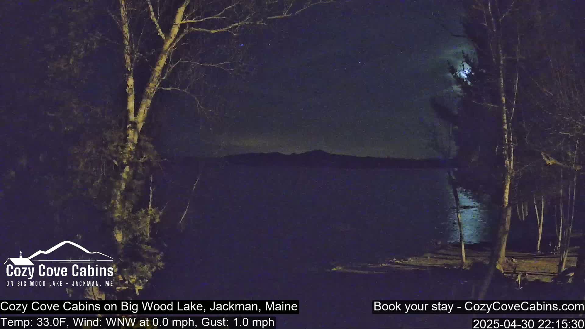 A nighttime view of a calm lake under a partly cloudy sky with a bright moon, seen from a lakeside vantage point among leafless trees.