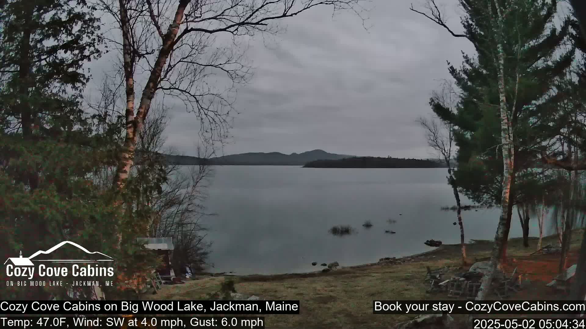 A calm lake with distant mountains is seen under an overcast sky, from a lakeside vantage point with bare trees and a small building visible in the foreground.