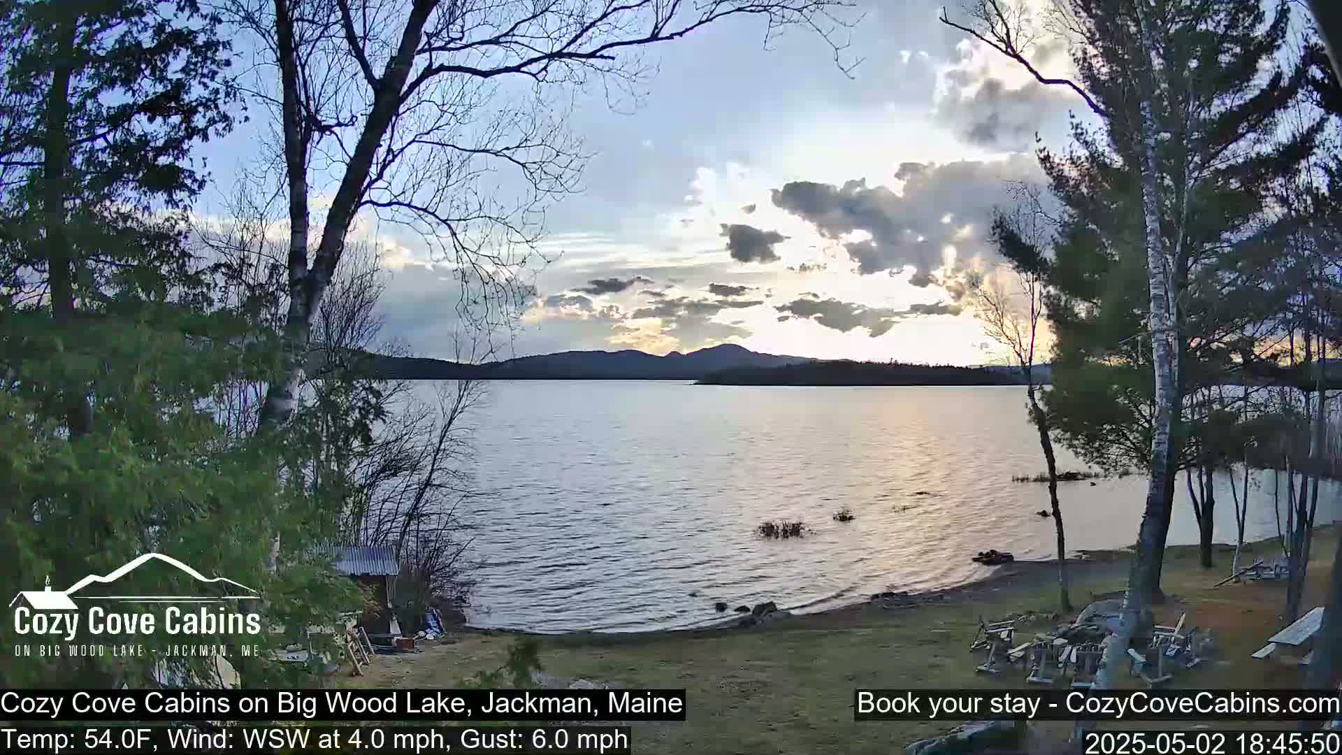 A calm lake at sunset is seen from a lakeside yard with trees and a fire pit, under a partly cloudy sky.