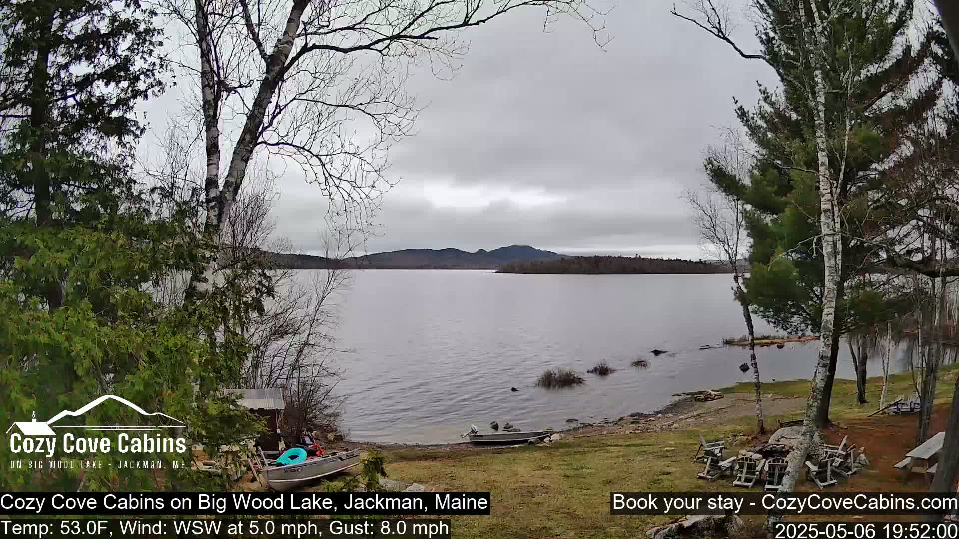A calm lake with a few boats on the shore, under an overcast sky, is seen from a lakeside property with bare trees and patchy grass.