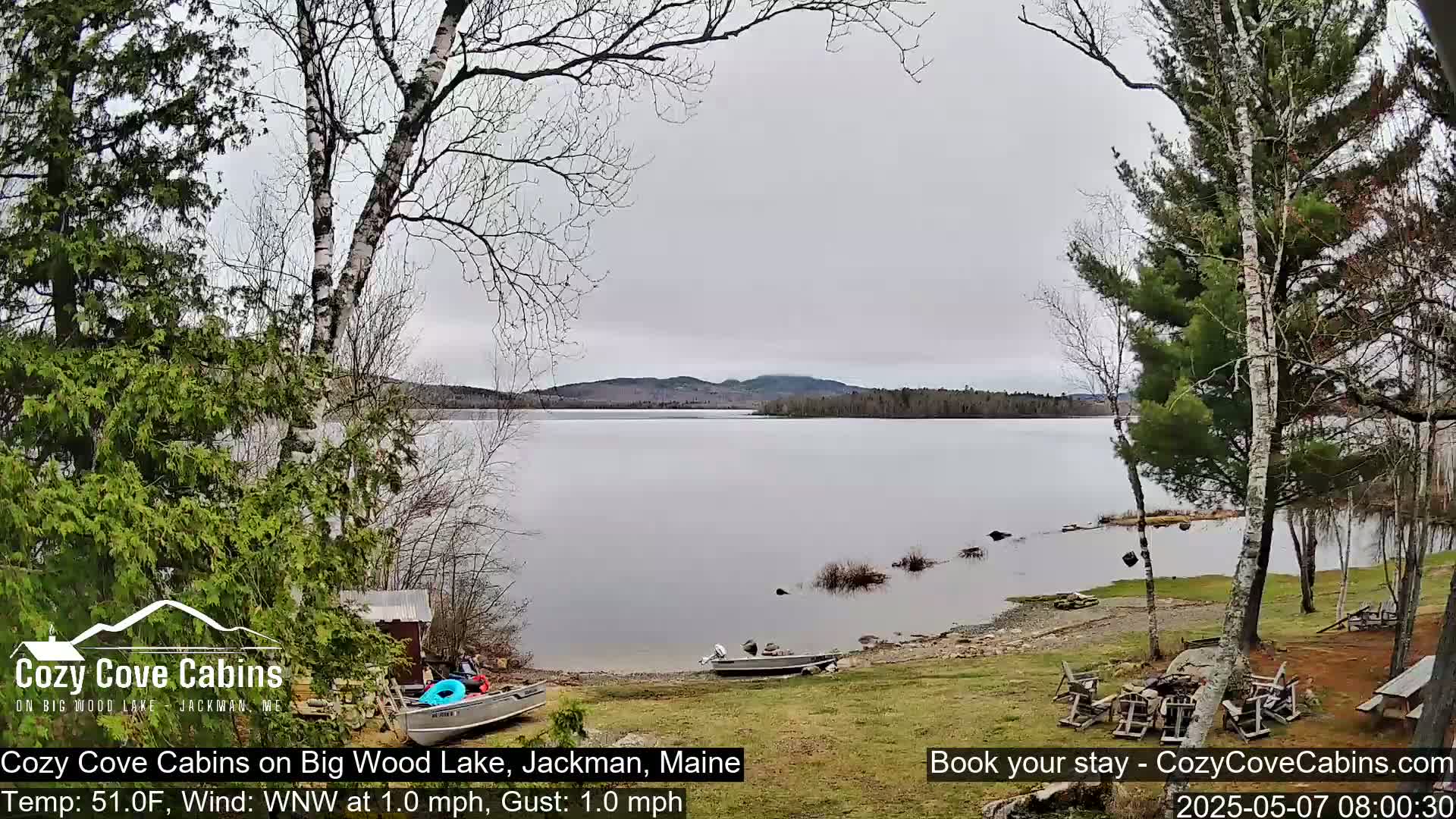 A calm lake under an overcast sky is seen from a lakeside property with several boats on the shore, surrounded by trees, some of which are budding.