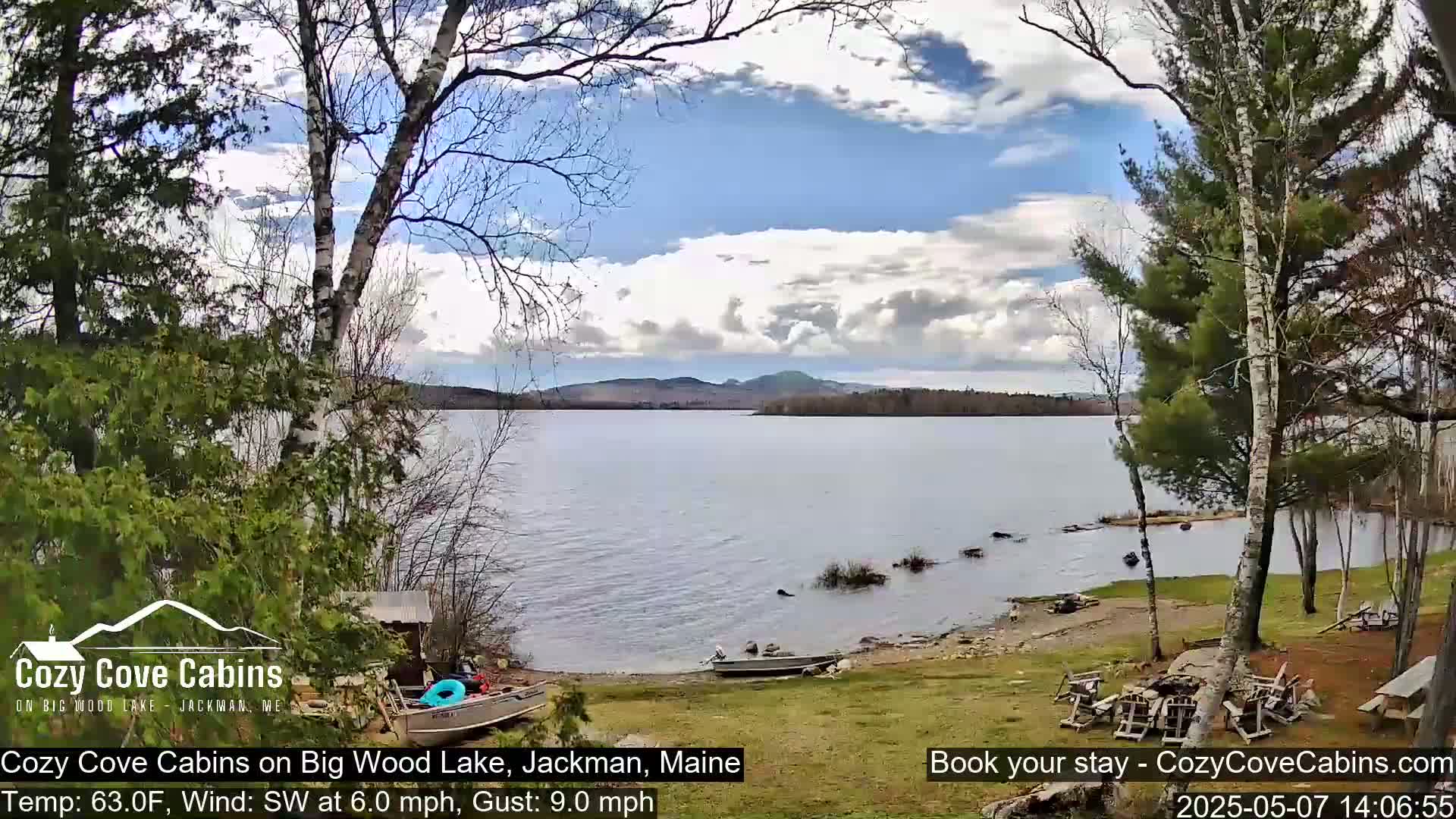 A partly cloudy day shows a calm lake with a small boat on a sandy shore, surrounded by trees and a grassy area with outdoor furniture.