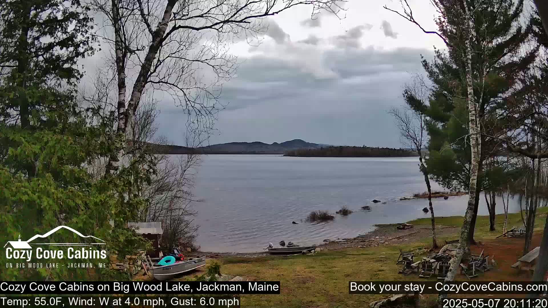 A mostly cloudy day reveals a calm lake with a shoreline featuring several small boats and a grassy area with sparse trees.