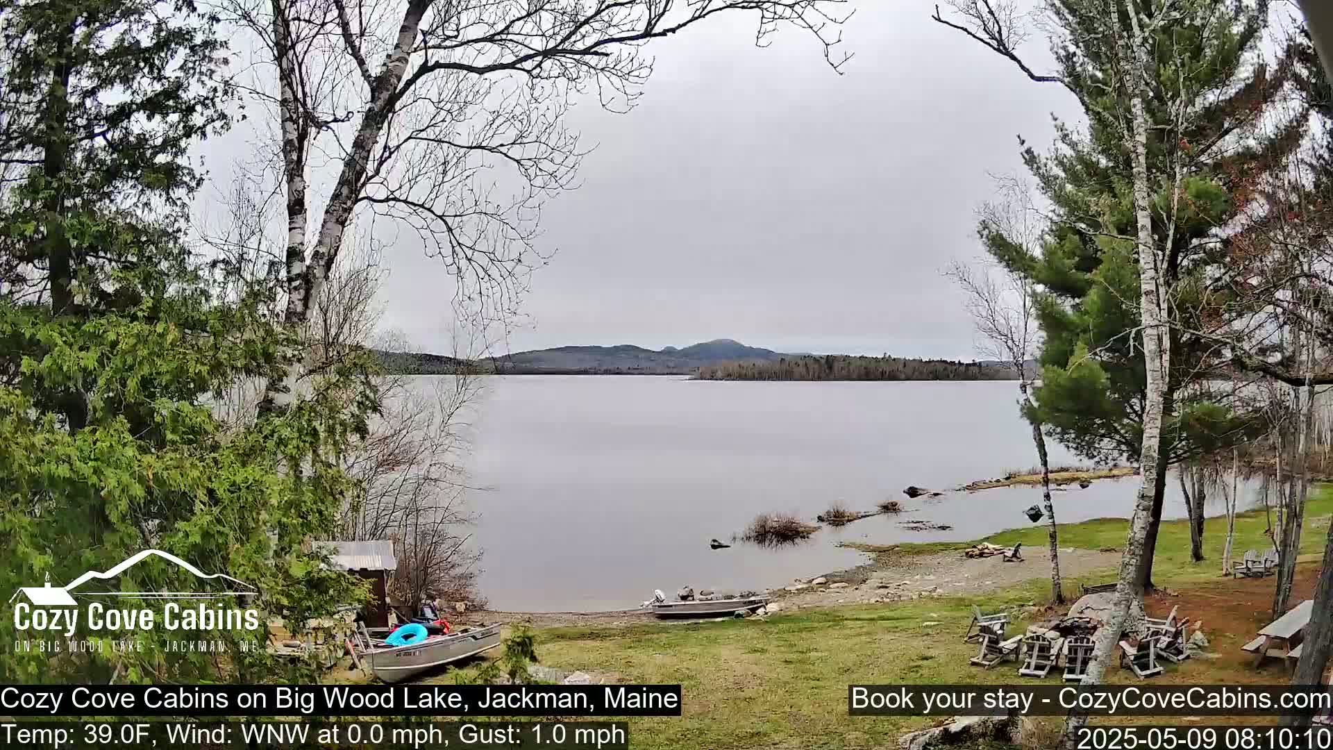 A calm, overcast day reveals a lake with a rocky shoreline, boats, and a grassy area with outdoor furniture, all nestled amongst trees.