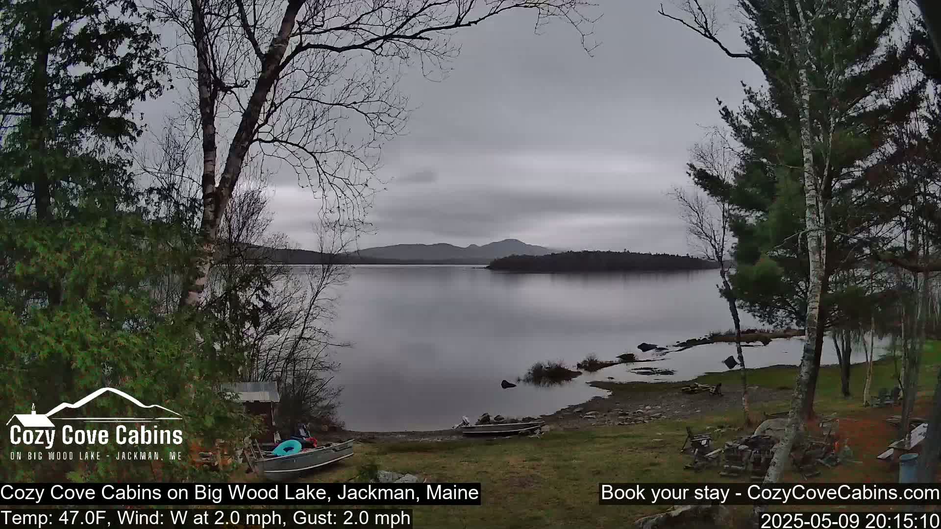 A calm, overcast day reveals a lake with a shoreline dotted with trees and a small boat, mountains visible in the distance.