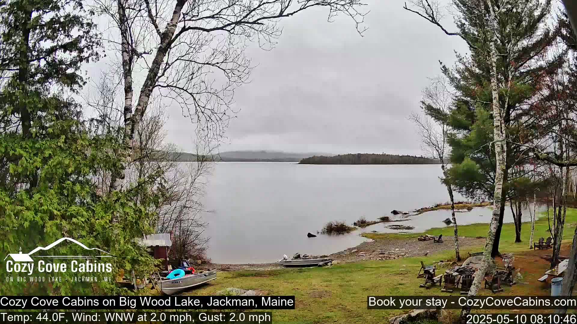 A calm lake with a rocky shoreline is viewed from a grassy area with trees and a couple of boats on the shore under an overcast sky.