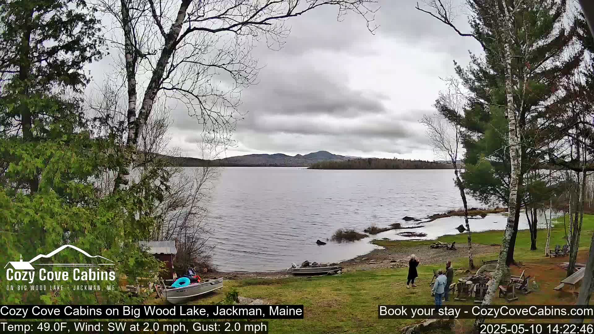 A calm lake under an overcast sky is viewed from a lakeside grassy area with several people standing near boats and outdoor furniture.