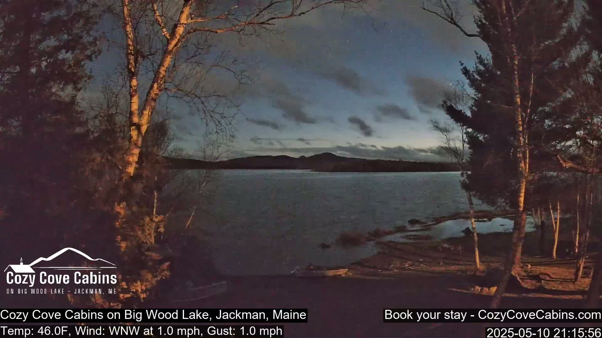 A calm lake at dusk is seen from a lakeshore, with trees silhouetted against a twilight sky.
