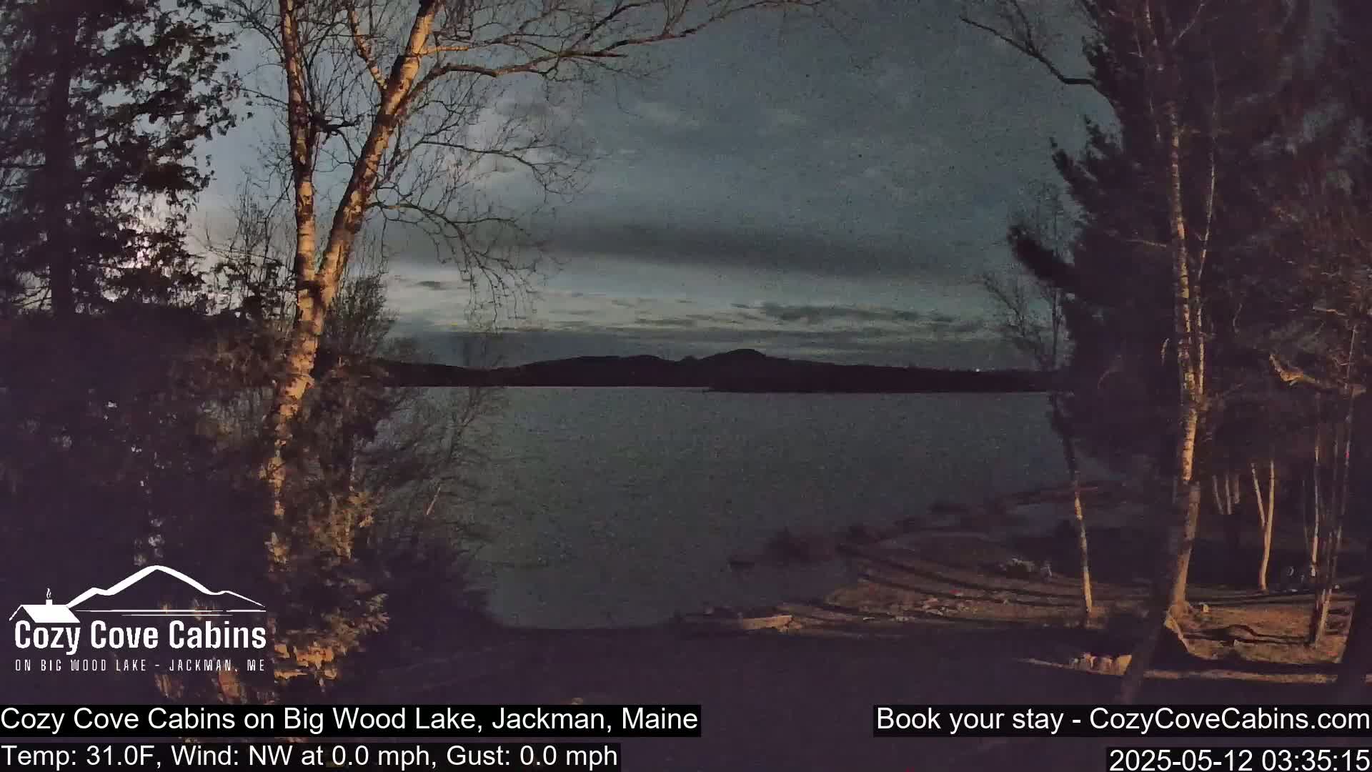 A dark, calm lake is seen at dusk from a wooded shoreline under a mostly cloudy sky.