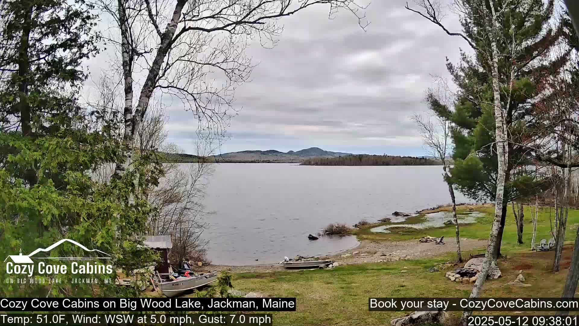 A calm lake with a rocky shoreline, partially-leafed trees, and two boats sits under a mostly cloudy sky.