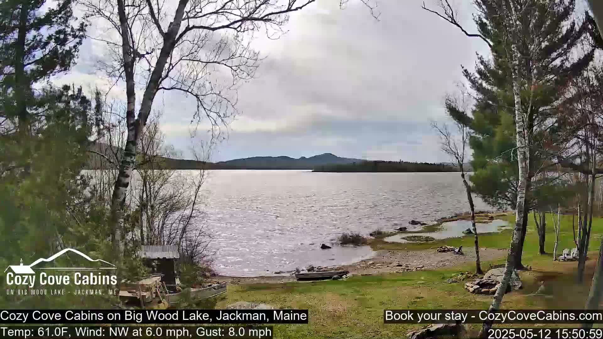 A mostly cloudy day shows a lake with a gently sloping shoreline, trees, and a small boat pulled ashore.