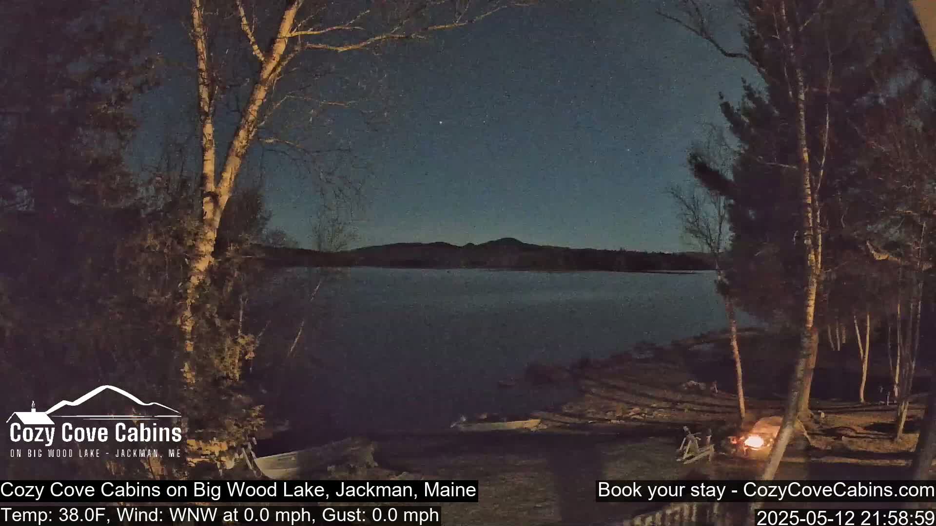 A calm, starry night scene shows a lake under a dark sky, with trees and a small fire visible in the foreground.