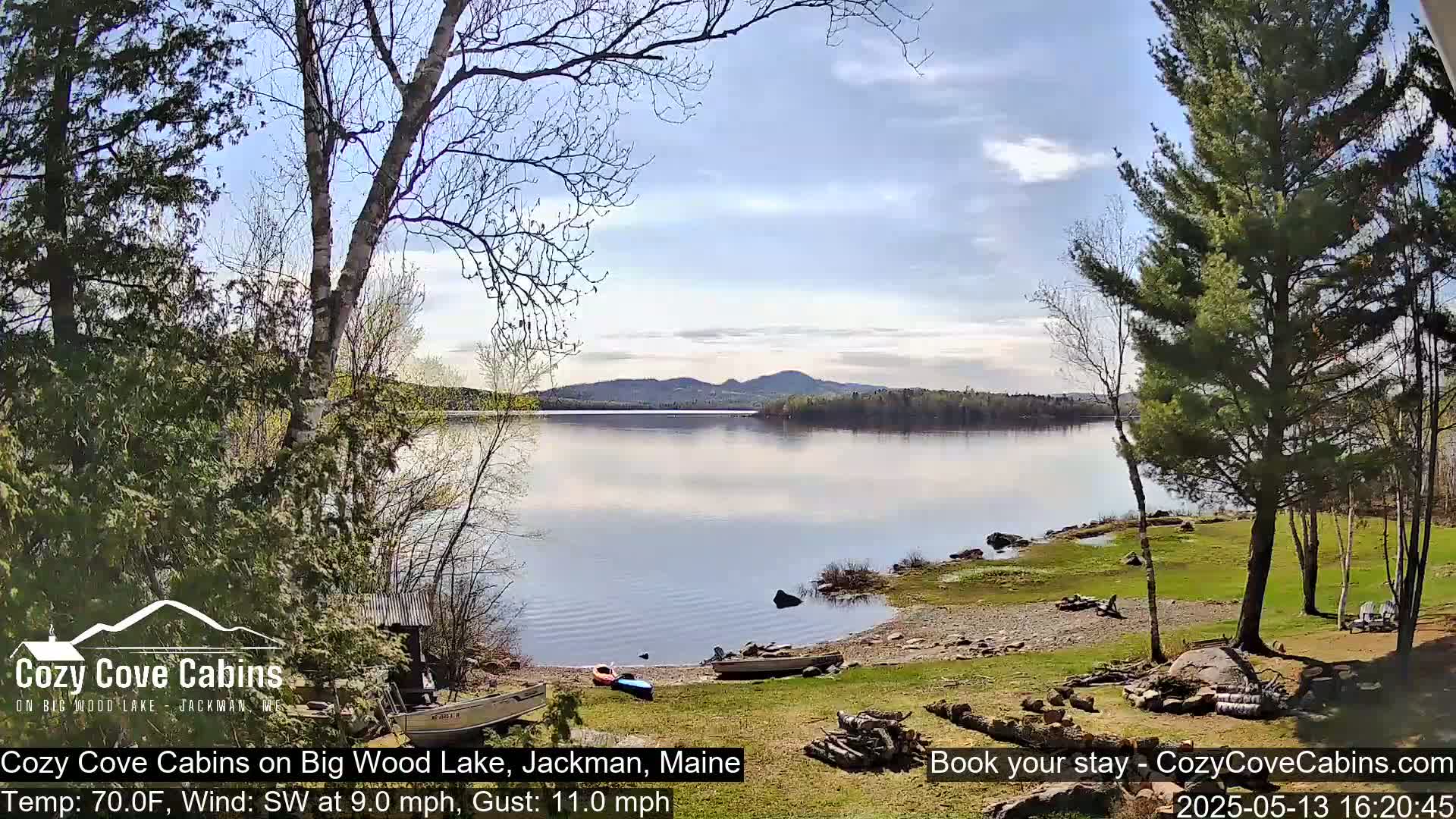 A calm lake with a sandy shore, boats, and trees under a partly sunny sky.