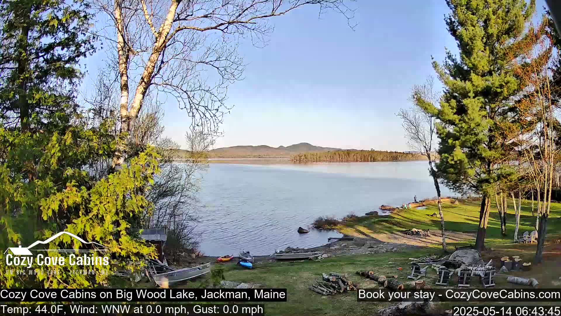 A calm lake with a shoreline featuring boats, trees, and a grassy area under a clear, sunny sky.