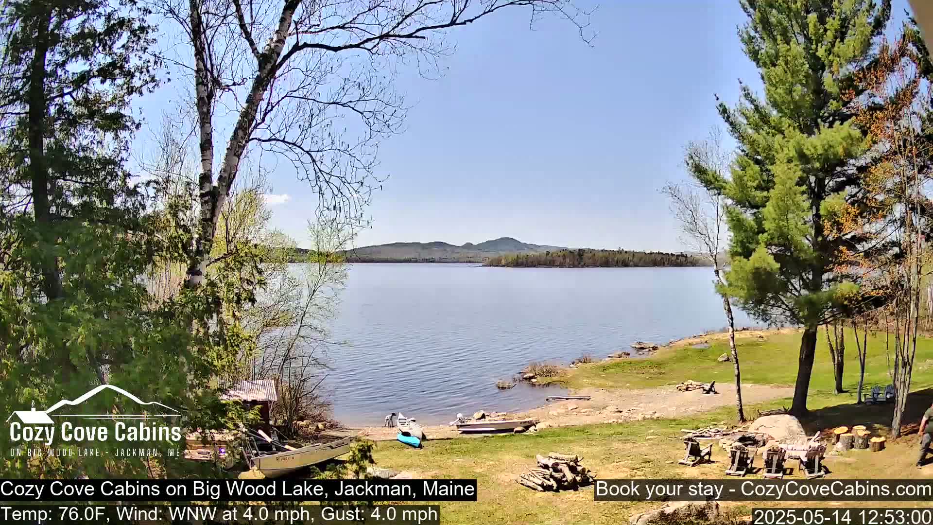 A tranquil lake scene on a sunny day with a gentle wind shows a sandy shoreline bordered by lush green grass and trees, with several small boats pulled ashore.