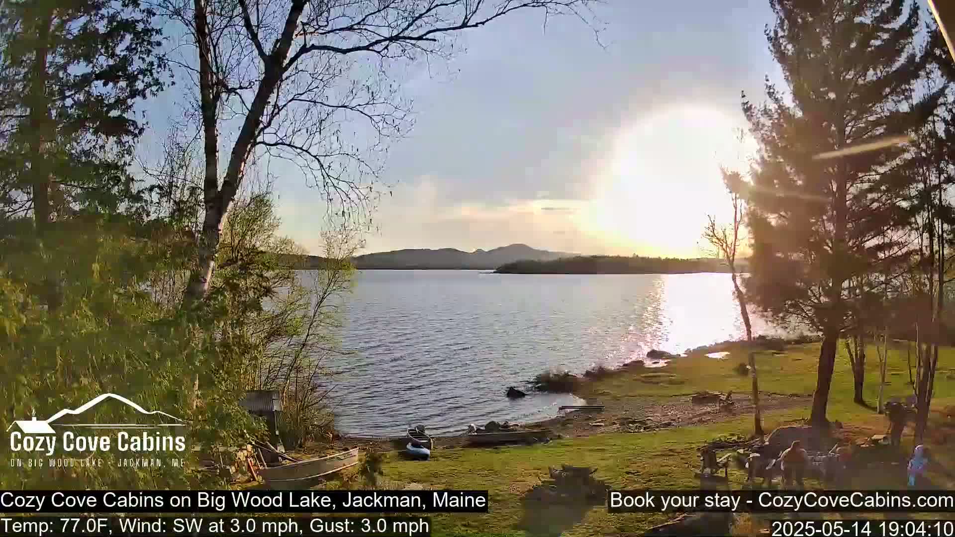 A calm lake at sunset, with a gentle southwest wind, is viewed from a lakeside grassy area with trees and several small boats.