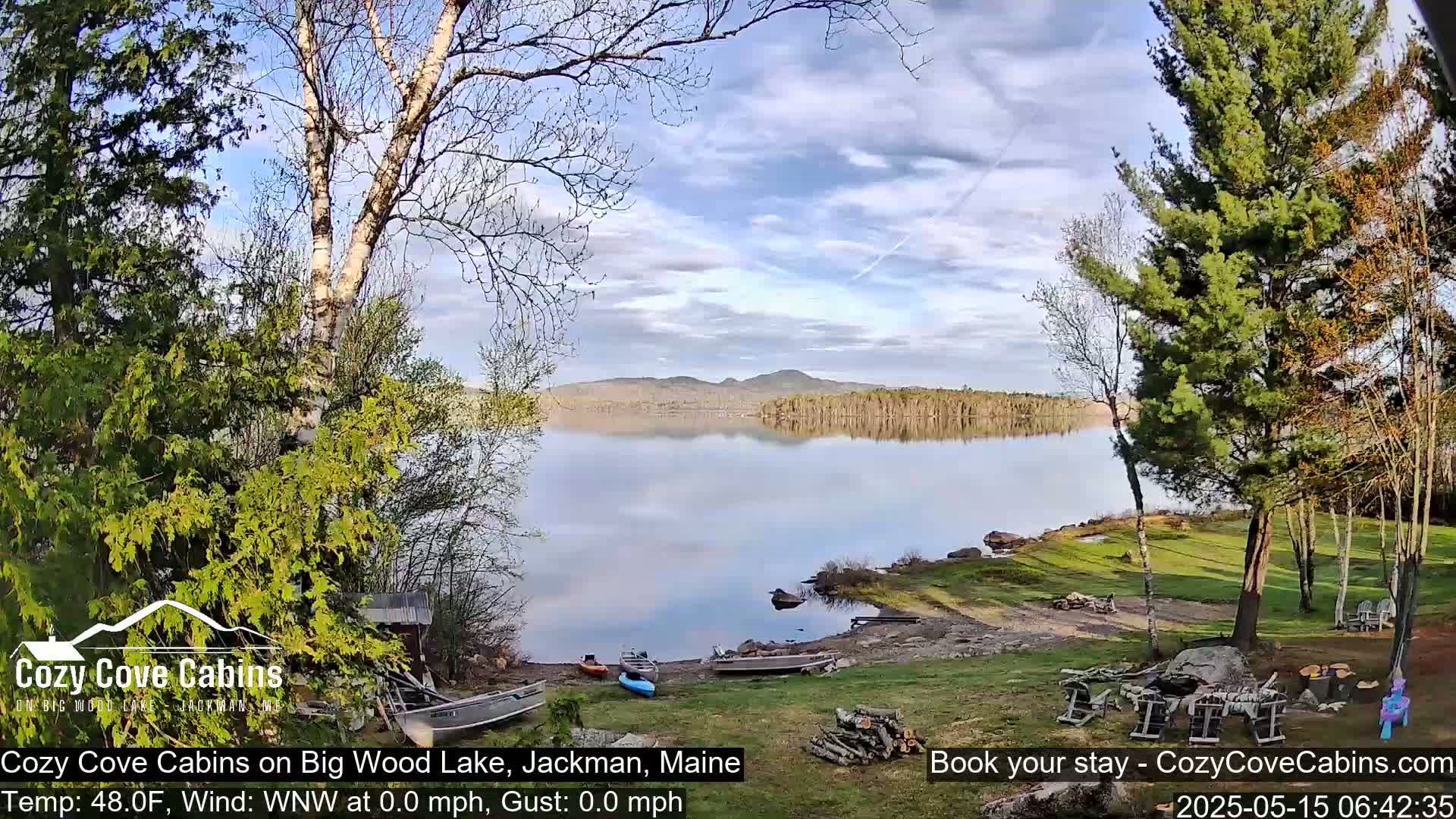 A calm lake with a few boats on its shore, surrounded by trees under a partly cloudy sky.