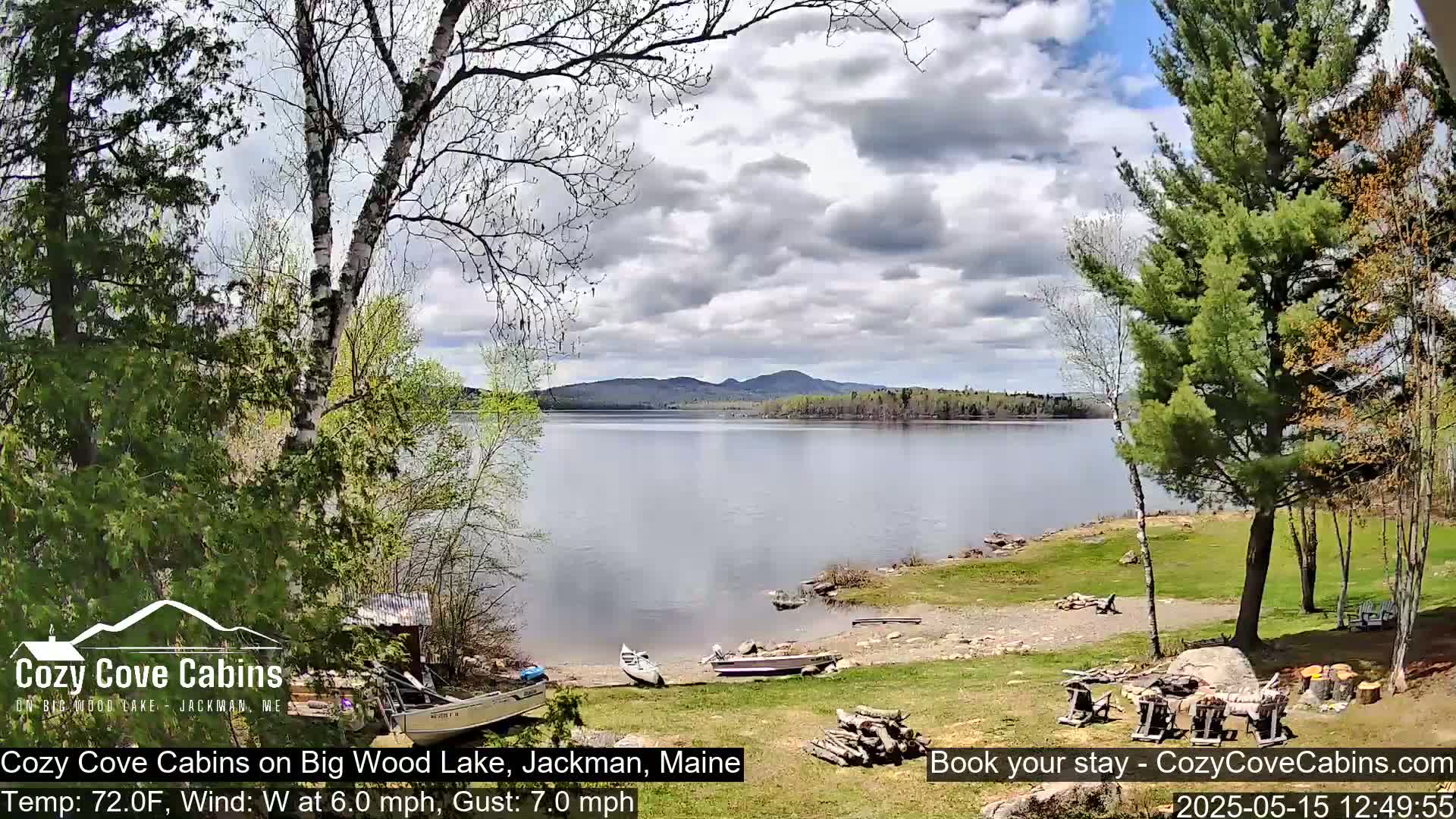 A calm lake is seen from a lakeside property under a partly cloudy sky, with boats on the shore and trees in the foreground.