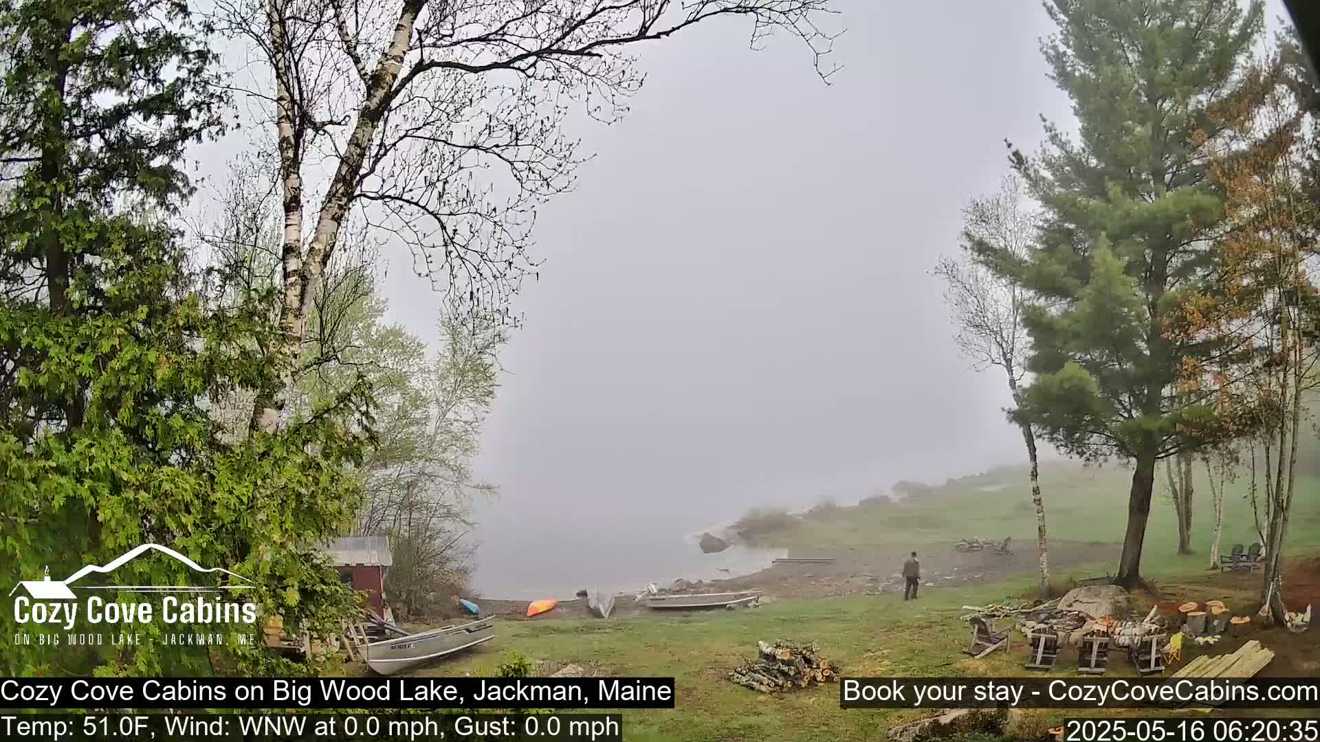 A foggy lakeside scene shows several boats and a person near a wooded area with a woodpile and cabins visible in the distance.