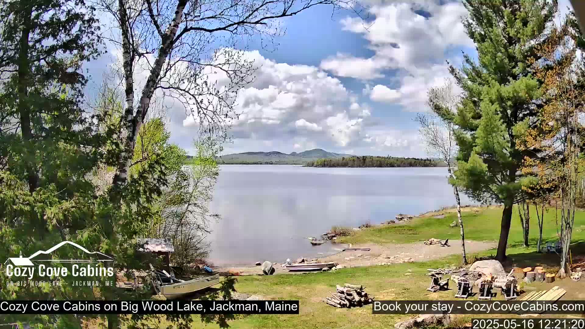 A tranquil lake scene under a partly cloudy sky, with trees lining a grassy shore where boats are beached near a woodpile.