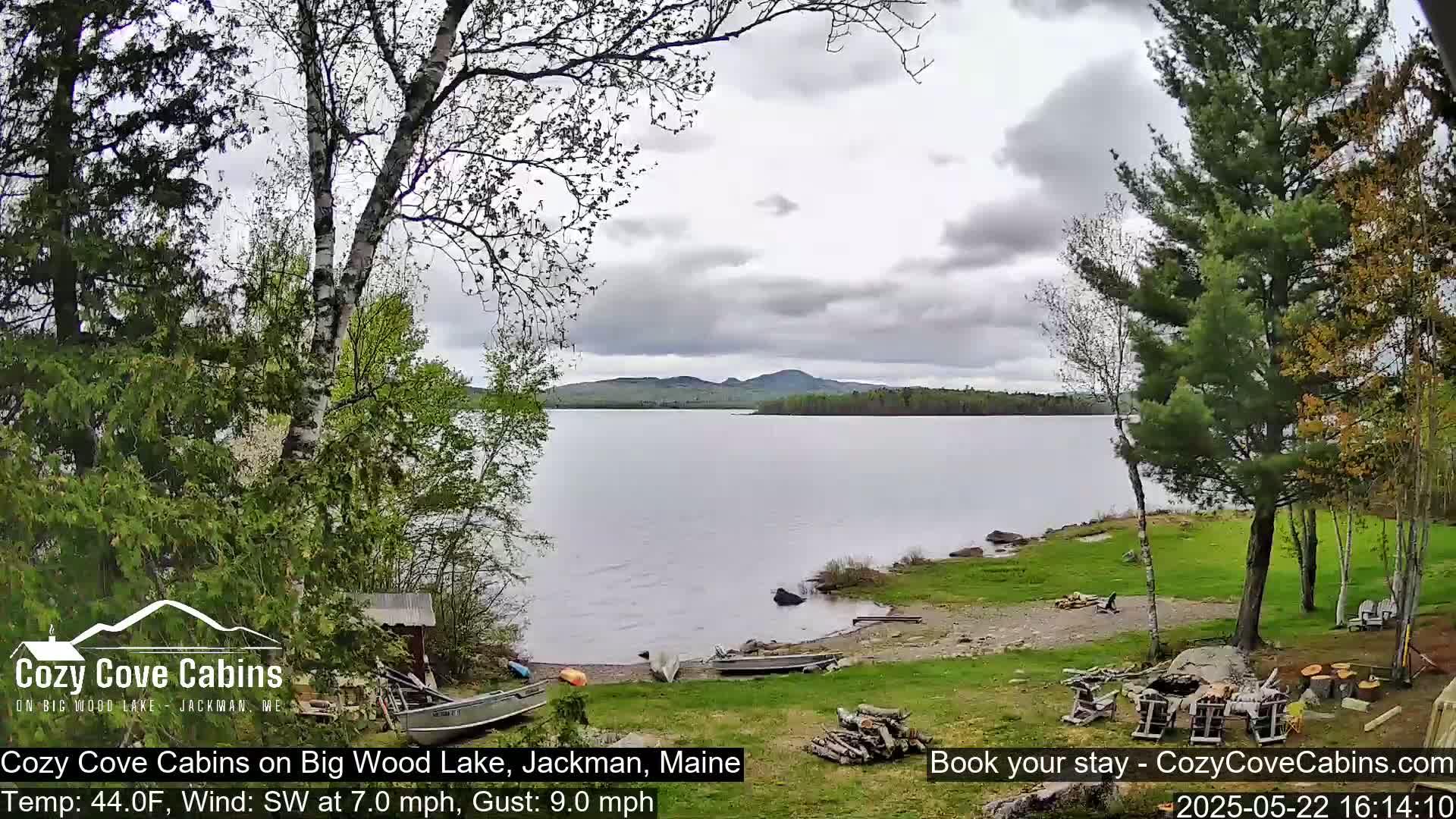 A calm lake with a shoreline featuring trees, grassy areas, and a few boats under an overcast sky with a southwest wind.