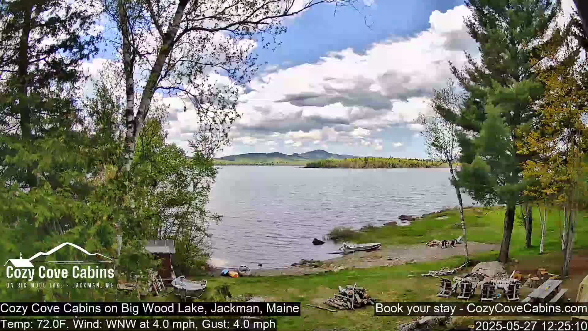 A partly cloudy day reveals a tranquil lake scene with a shoreline featuring trees, a small sandy beach area with some outdoor furniture, and distant mountains under a mostly sunny sky.