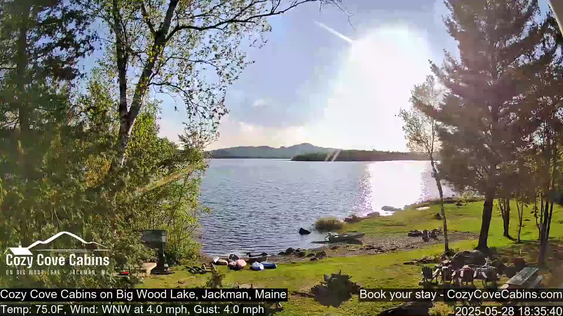 A sunny day with a light wind shows a tranquil lake scene, featuring several canoes pulled ashore near a lakeside cabin area.