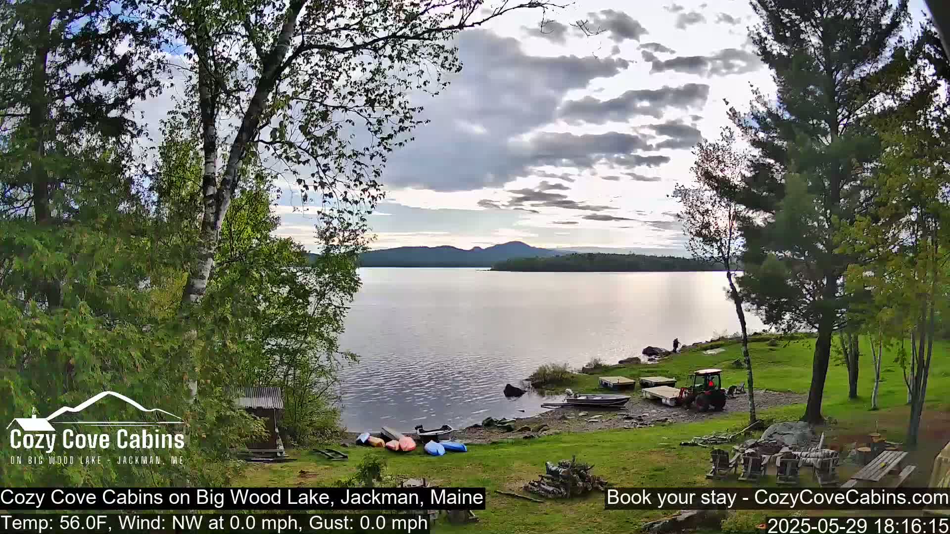 A calm lake with a few boats on the shore, surrounded by green trees under a partly cloudy sky.