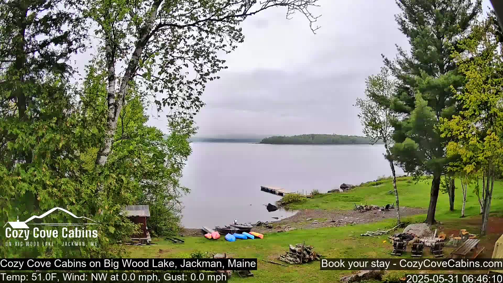 Kayaks and a small boat are on the shore of a calm lake, under an overcast sky, surrounded by green trees and a small cabin.