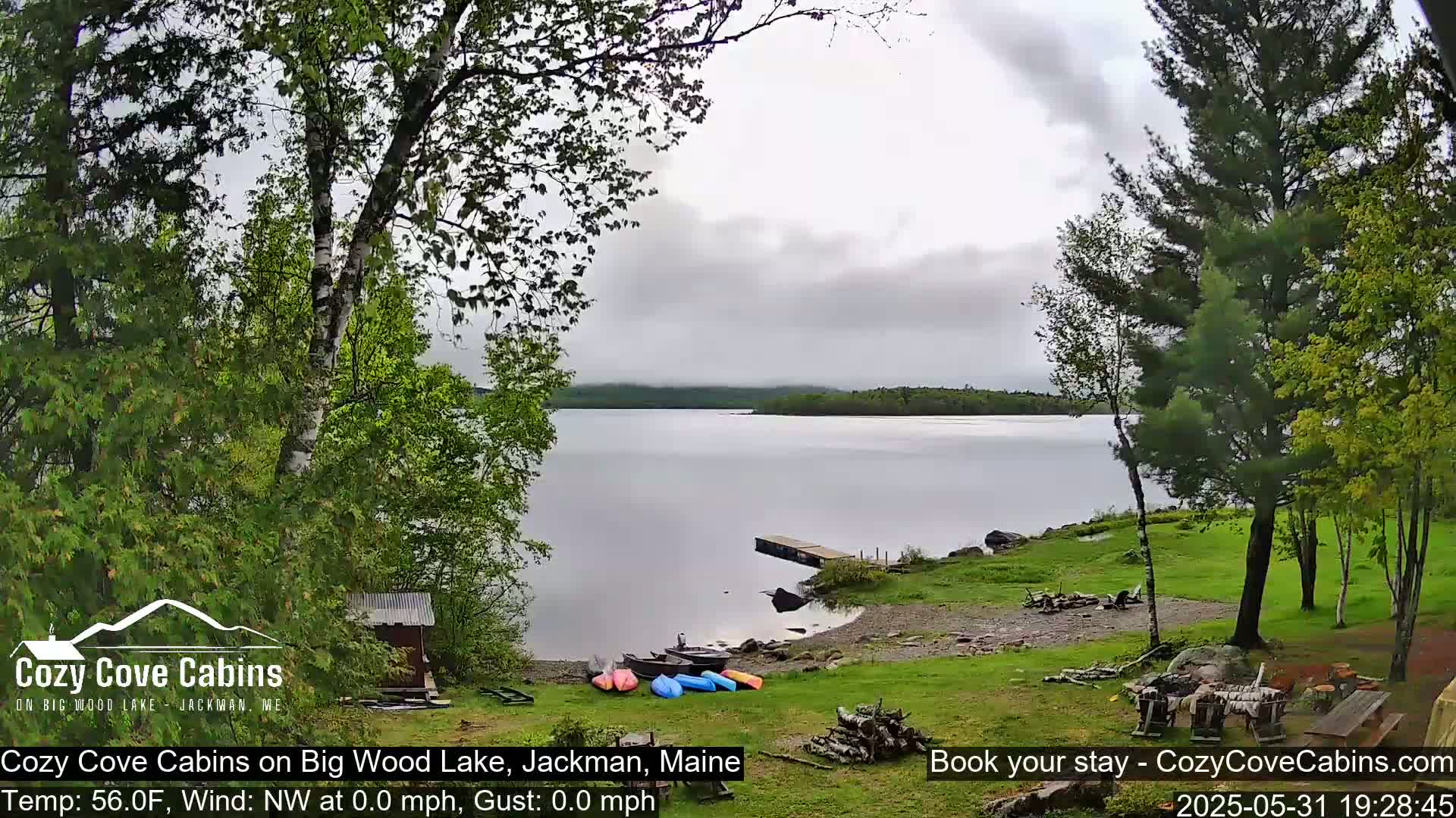 A calm, overcast day on a lake, viewed from a lakeside grassy area with trees, a small dock, and several kayaks pulled ashore.
