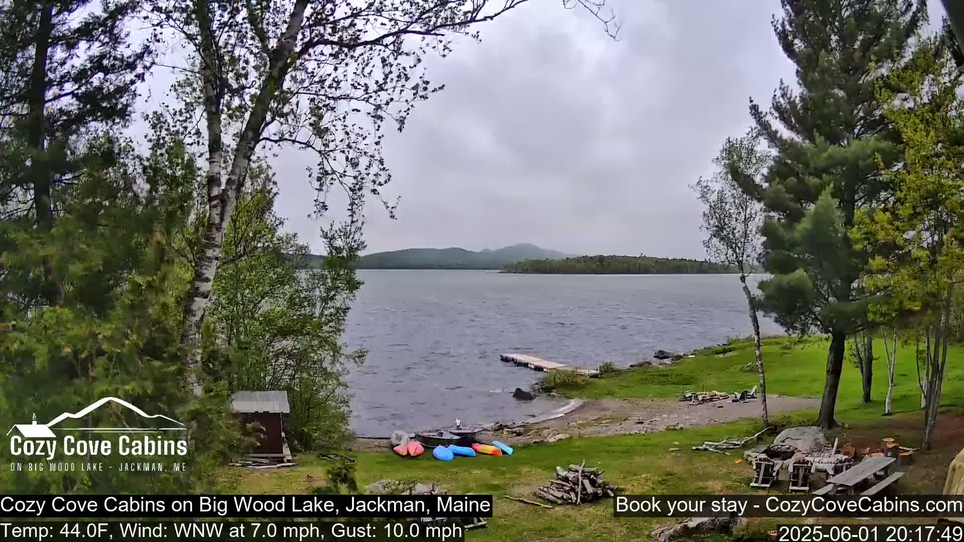 Kayaks are on the lakeshore near a small shed and picnic table, with a windy, overcast day over a lake and distant forested hills.