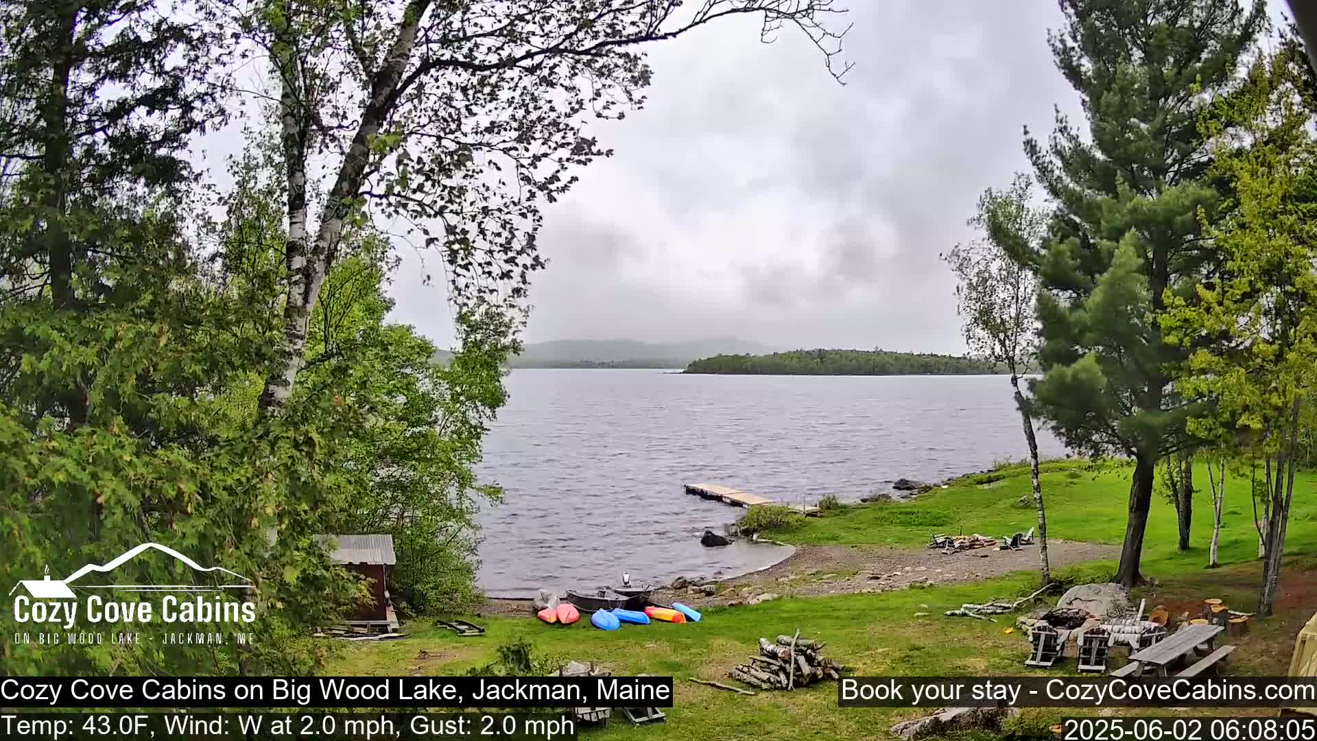 Kayaks are on the shore of a calm lake under an overcast sky, surrounded by green trees.