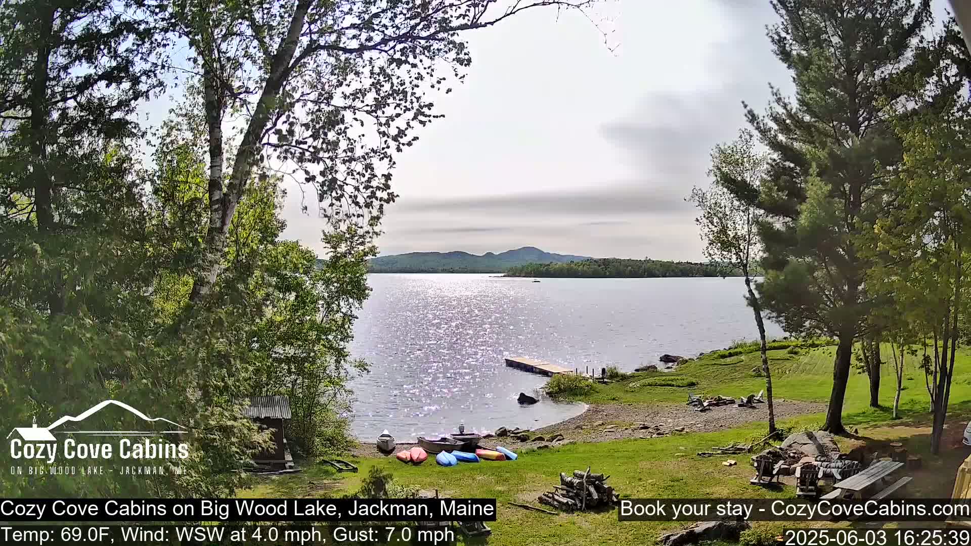 Kayaks are lined up on a grassy lakeshore with a small dock extending into a sparkling lake under a partly cloudy sky, with trees in the foreground and background.