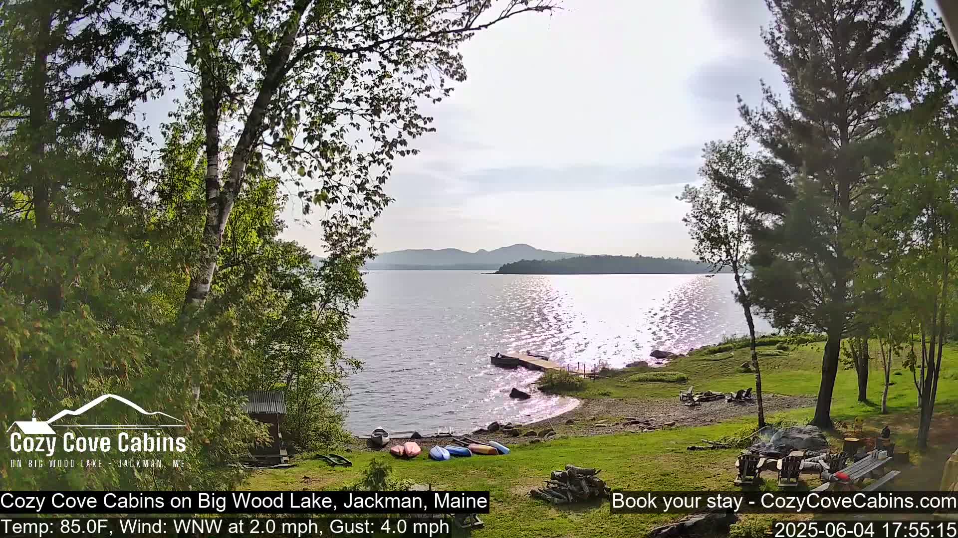 A lakeside view on a partly sunny day with a gentle westerly wind shows calm water reflecting sunlight, a sandy shoreline with kayaks and a woodpile, and evergreen trees bordering the property.