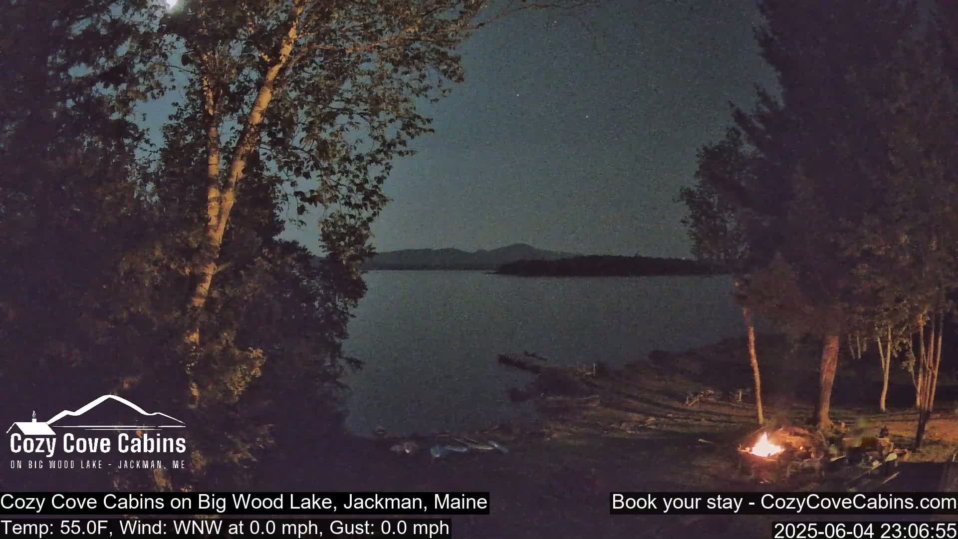 A nighttime scene shows a calm lake under a mostly clear, dark sky with a campfire burning on the lakeshore near some trees.