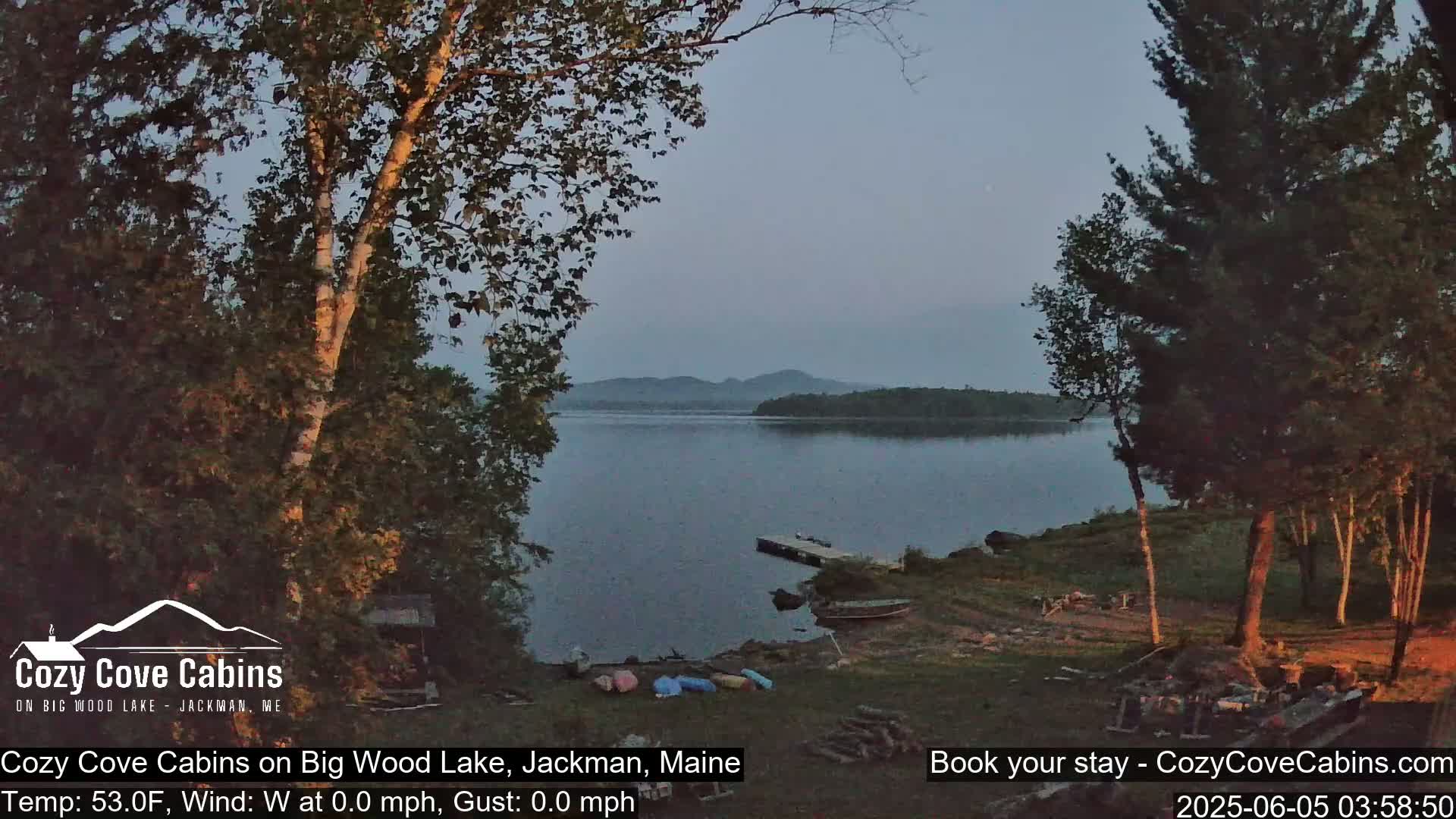 A calm lake at dusk is seen from a lakeside property, with several canoes and a small dock visible in the foreground, surrounded by trees under a clear, twilight sky.