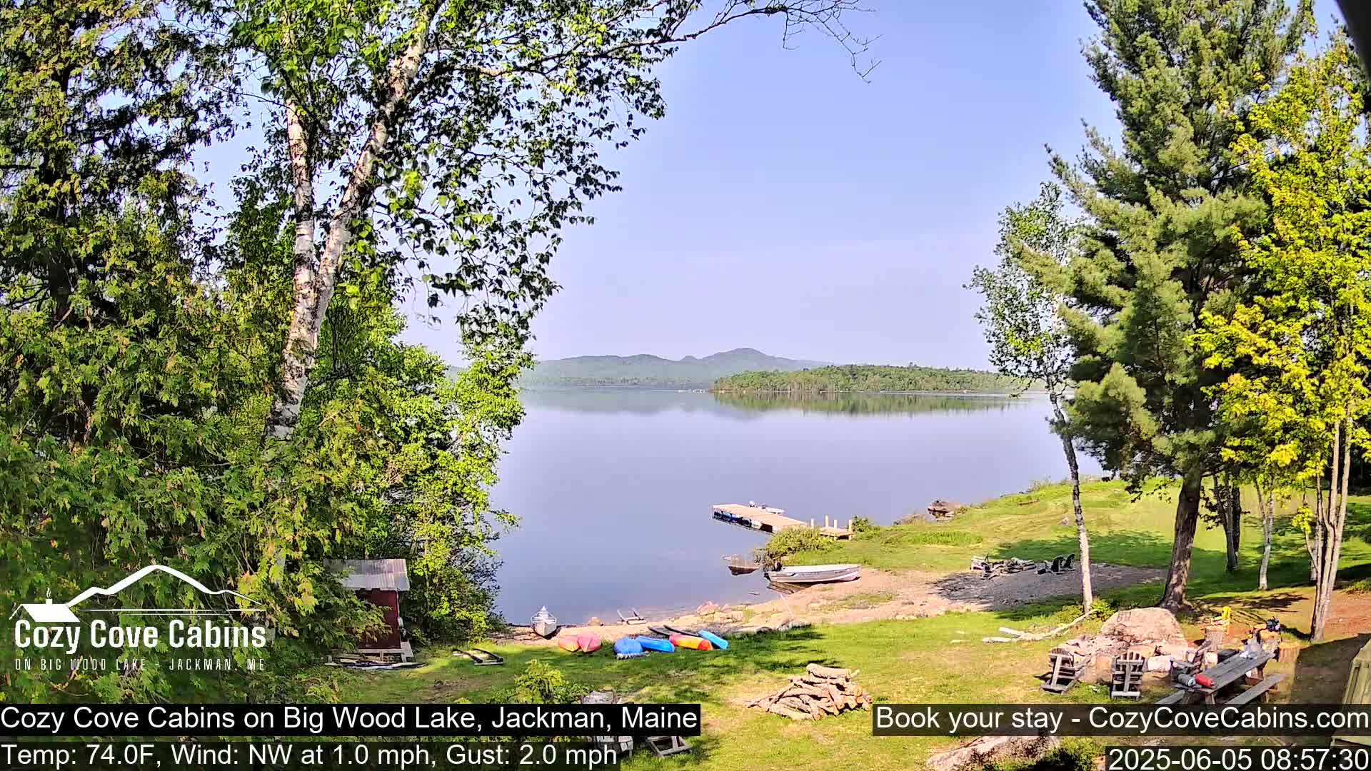 A calm lake with a shoreline featuring kayaks, a small boat, and a dock is surrounded by lush green trees under a clear blue sky.