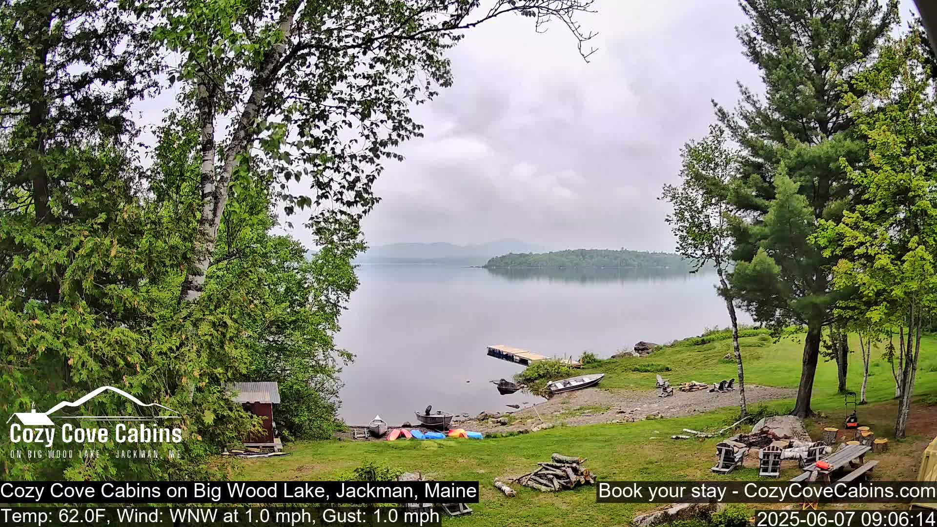 A calm lake with a small dock and several boats sits beside a grassy area with trees and a fire pit under an overcast sky.