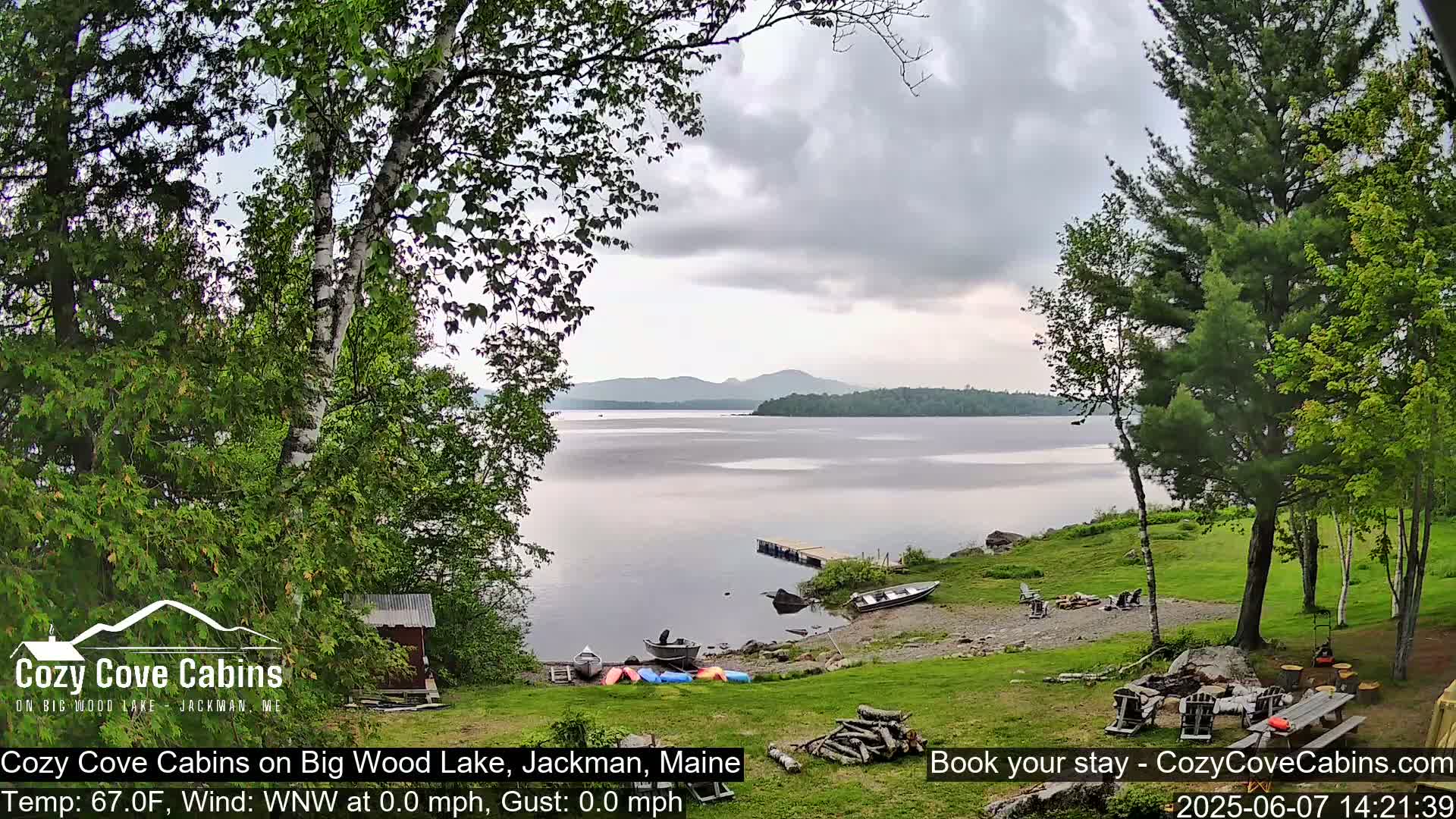 A calm lake with a cloudy sky is seen from a lakeside yard with several boats pulled ashore near a fire pit and picnic table.