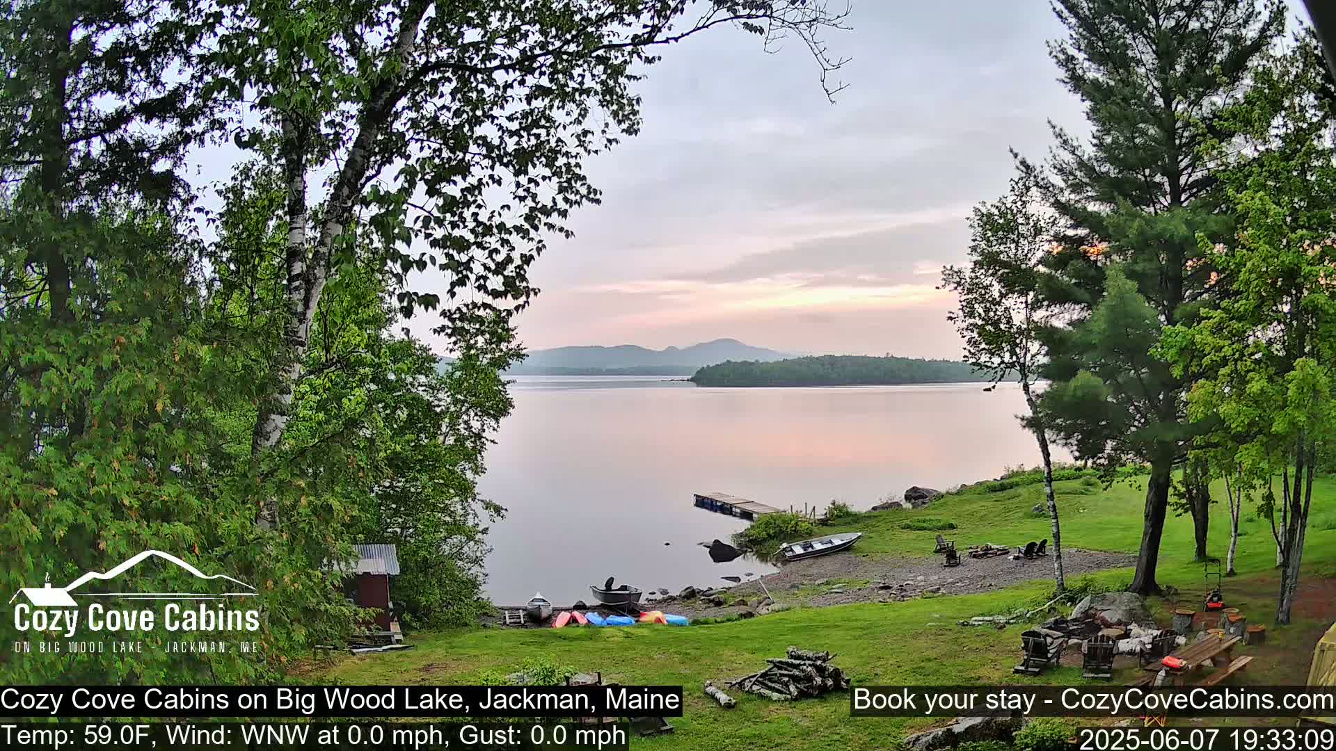 A calm lake at sunset is viewed from a grassy lakeshore with several boats and canoes pulled ashore near a small cabin, surrounded by lush green trees.