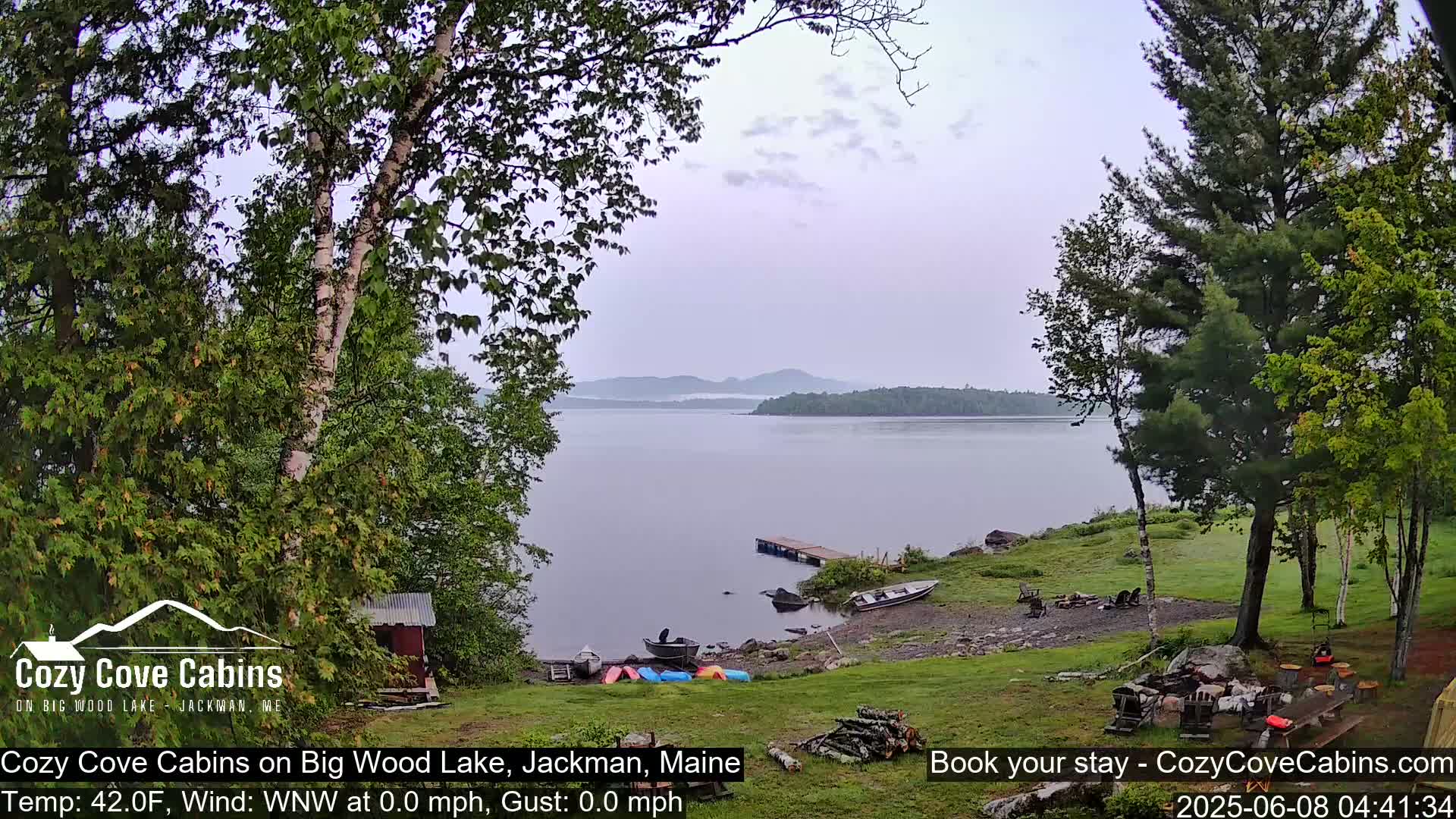 A calm lake with a few boats and a dock is seen from a lakeside property on an overcast day.