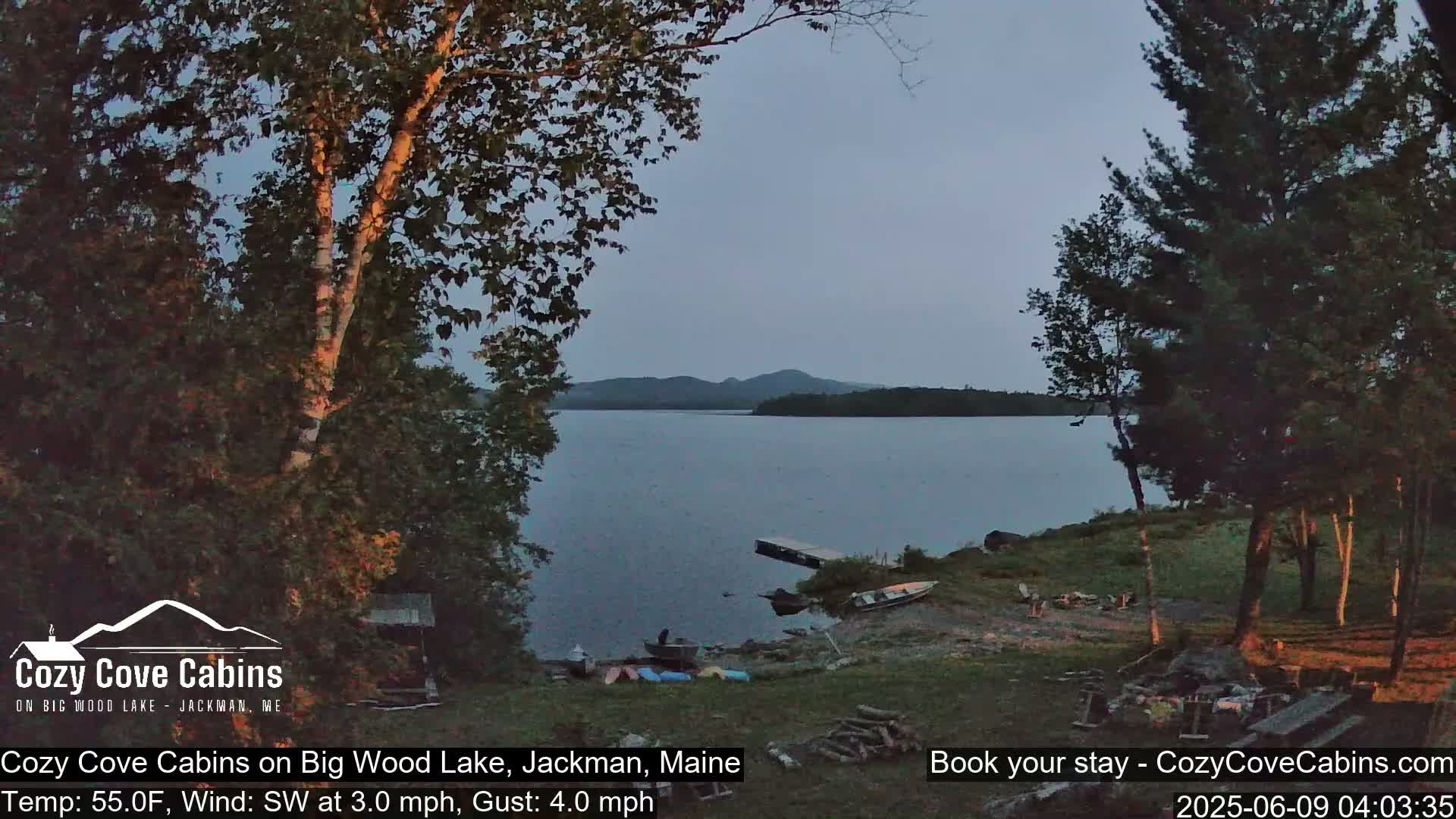 A tranquil lake scene at dusk, with calm water, a few boats on shore, and trees surrounding a grassy area.